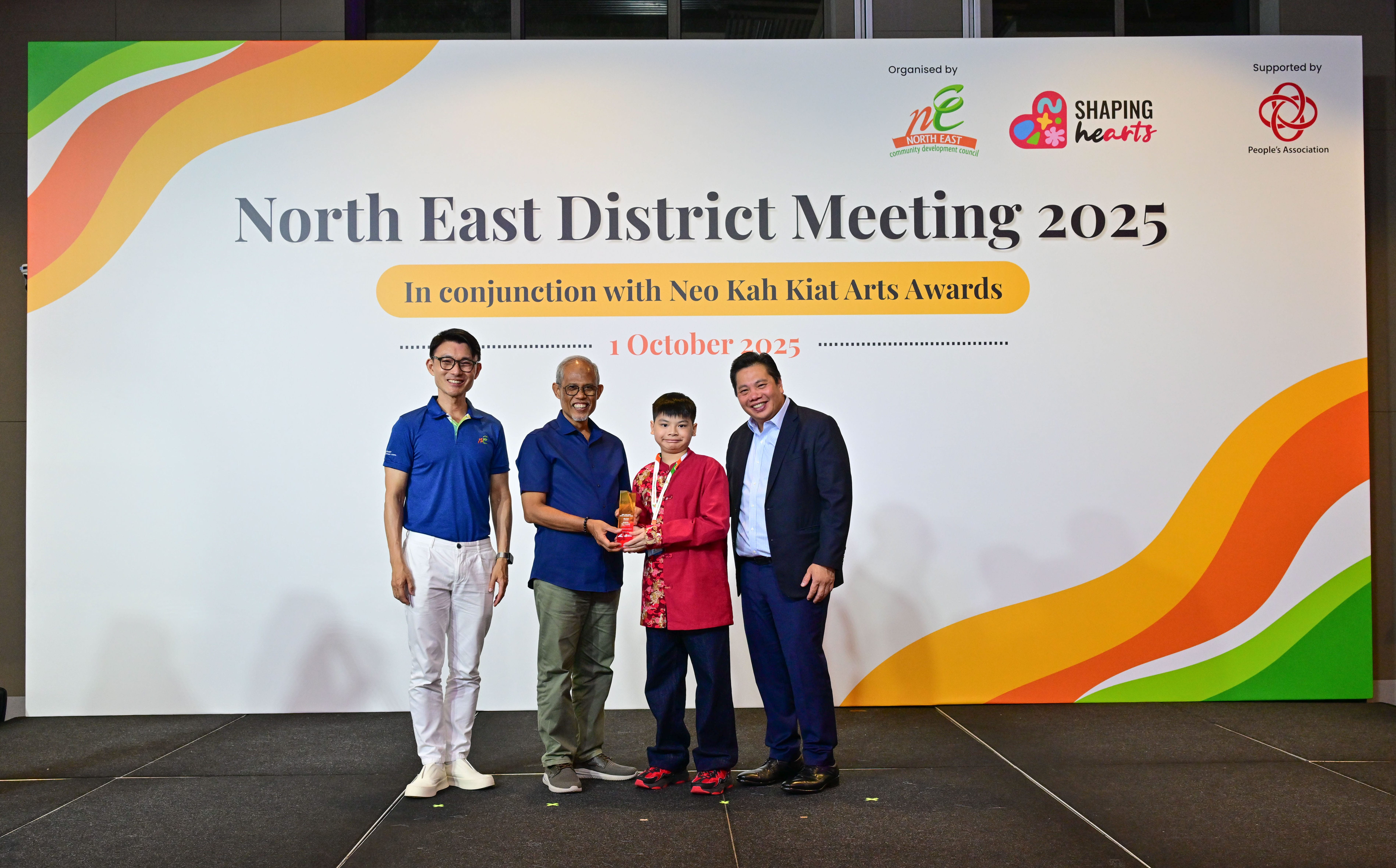 Award recipient standing on stage with the presenter, holding a acrylic plaque and posing for a group photo during the award ceremony, with a backdrop reading ‘North East District Meeting 2025 In conjunction with Neo Kah Kiat Arts Awards'